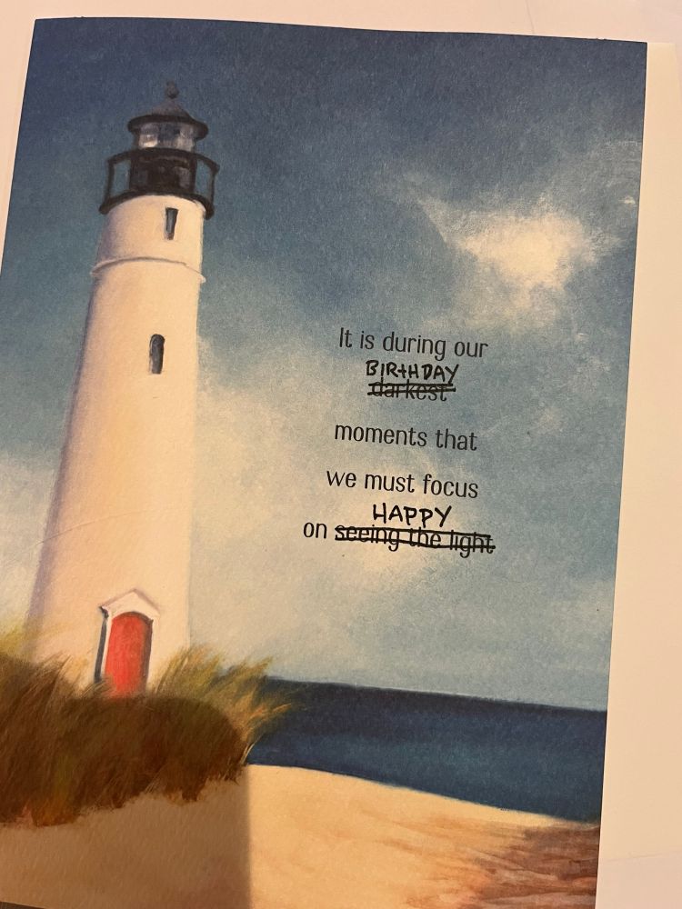front cover of a card with a painted lighthouse on it reading “It is during our (darkest) (which has been crossed out and replaced with “birthday”) moments that we must focus on (seeing the light) (which has been crossed out and replaced with “happy”).”