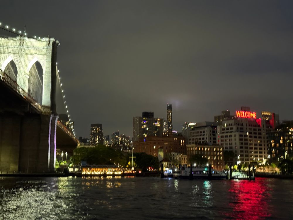 The Brooklyn Bridge and the Brooklyn skyline south of the bridge at night, viewed from a boat. One of the buildings has a neon sign that says “Welcome”
