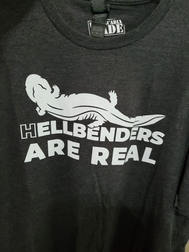 Black t shirt with white lettering saying " Hellbenders are real". There is an illustration of a hellbender amphibian above.