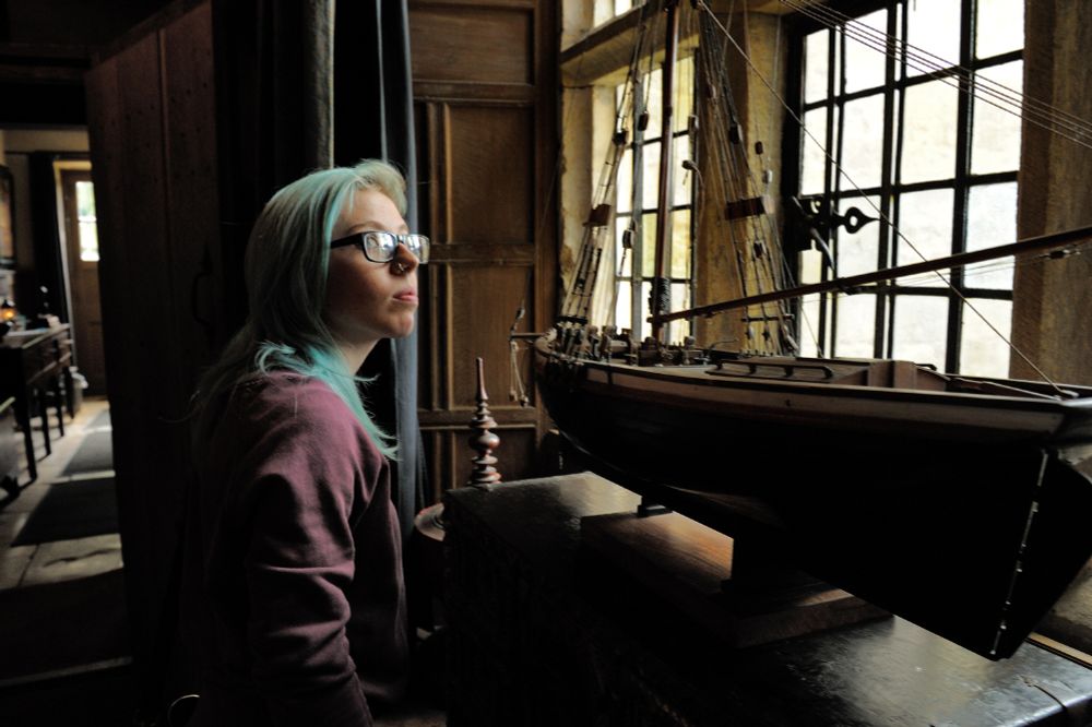 A young person with long blue hair, a maroon jacket and glasses studying a model boat at Snowshill Manor.