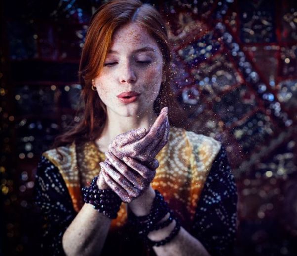 A redhead Caucasian woman blows glitter out of her hands.