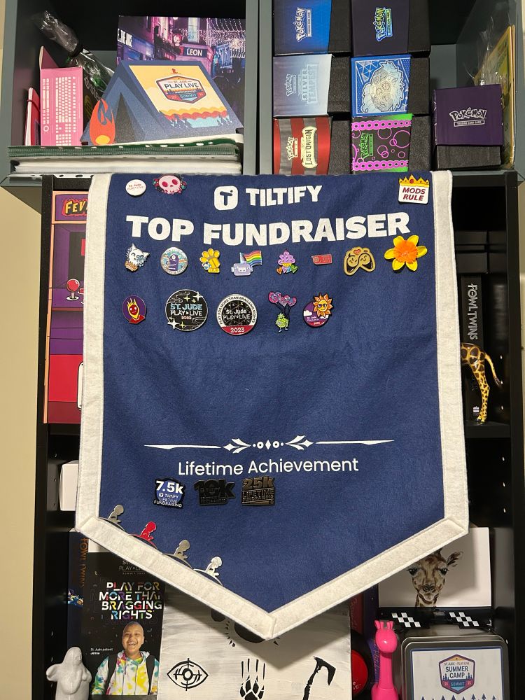 The photo shows a blue banner with a white edge. In the top middle reads “Tiltify Top Fundraiser” and blow it shows 13 enamel pin badges spread out the banner from left to right. Bottom middle reads “lifetime achievement” with 3 lifetime achievement badges that read 7.5k lifetime raised, 10k lifetime raised, 25k lifetime raised. There are also 4 magnet pins of the St. Jude Children’s Research Hospital logo. 