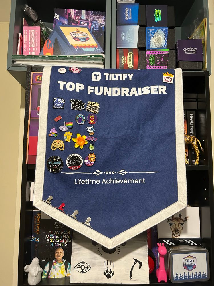 The photo shows a blue banner with a white edge. In the top middle reads “Tiltify Top Fundraiser” and blow it shows 16 enamel pin badges down the left side. Bottom middle reads “lifetime achievement” and shows 4 magnet pins of the St. Jude Children’s Research Hospital logo 