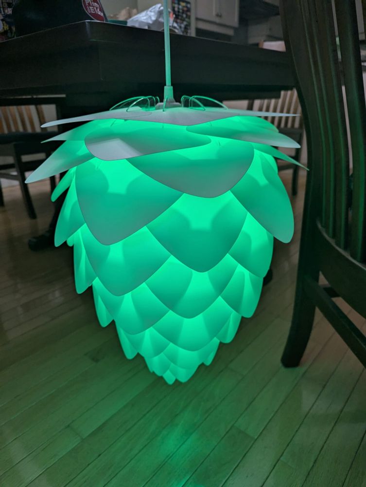 White light shade that looks like a pine or hop cone suspended between two chairs and illuminated with a green light