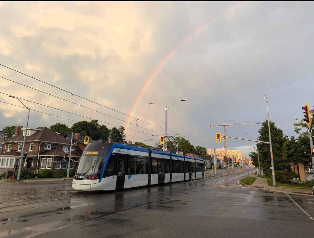 Blue & white ION streetcar with a rainbow in the sky behind it, going through the Union St intersection with visibly wet pavement