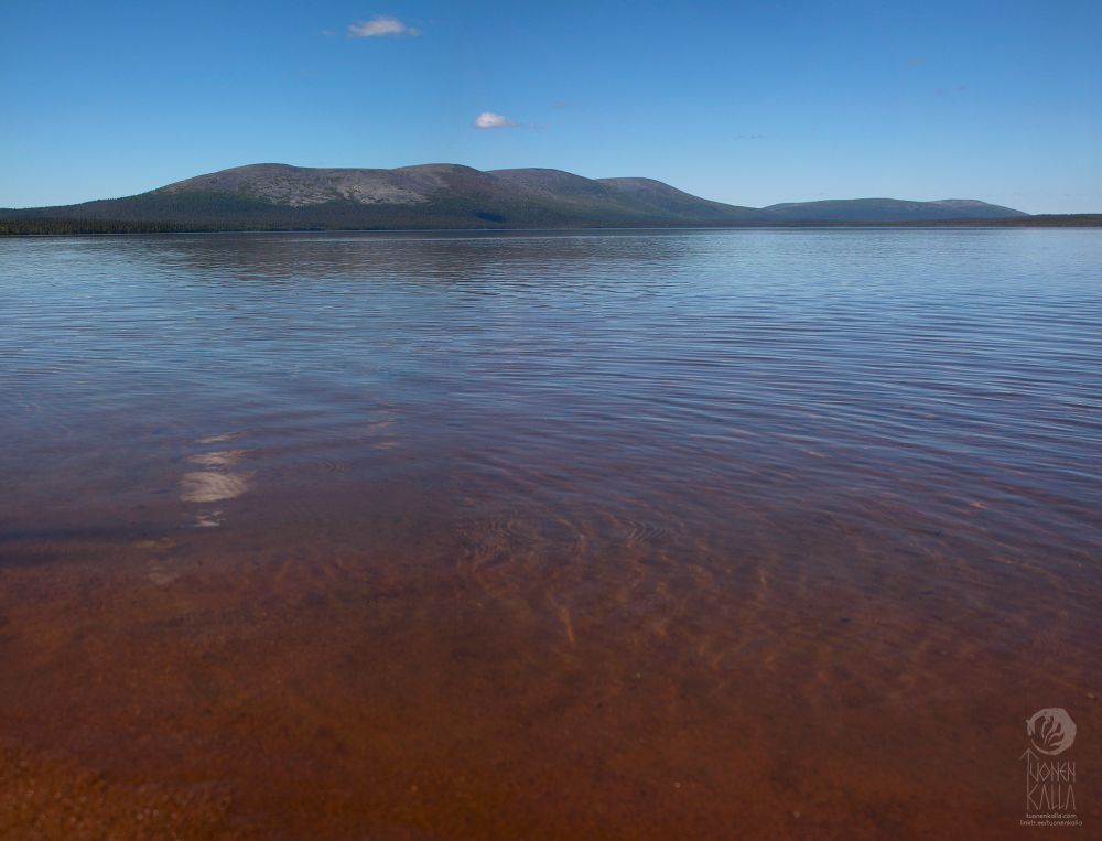 Photography of a sunny beach, there are fells visible on the other side of the lake. The sand is a shade of red.