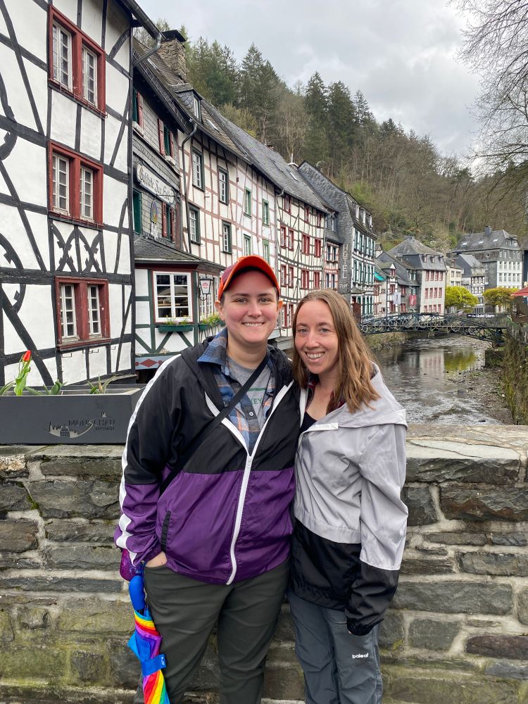 My wife and I posing for a photo in Monschau, Germany. I am wearing a VT Hokies hat, purple and black windbreaker, and green pants. My wife is wearing a grey and black jacket with grey pants. We are standing in front of a stone bridge over a creek with Bavarian-style houses in the background.