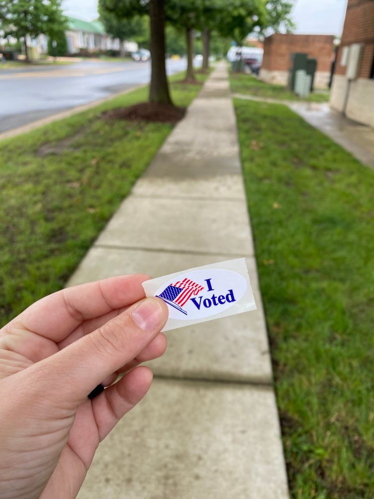 A hand holds an “I Voted” oval sticker with the American flag. The sticker is held up in front of a sidewalk with green grass on either side. There are trees and a road blurred in the background. 
