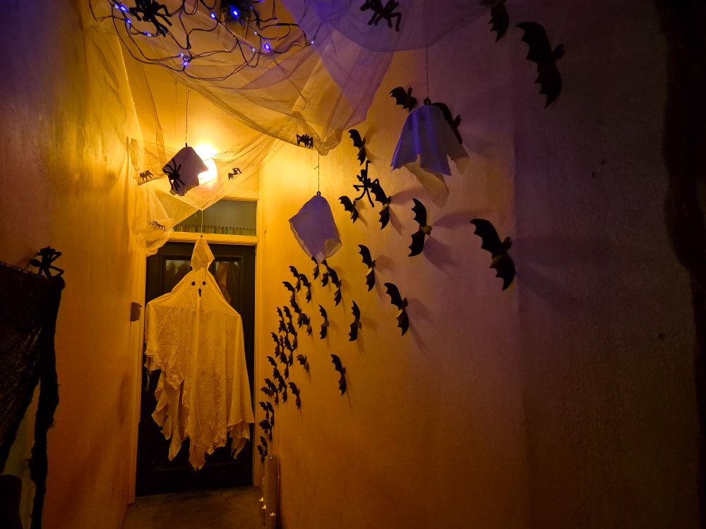 A recessed doorway. Grey tuille fabric is suspend from the ceiling above the door coming down the length of the passageway. A ghost made of a white sheet in hung in front of the door and card bats are swooping up the wall from the bottom right of the door. Pipe cleaner spiders and small ghosts are hung from the tuille. On the right of the passage are cardboard cylinders with electric tealights in the top of them to simulate candles. At the very top of the picture are some purple L.E.D lights.