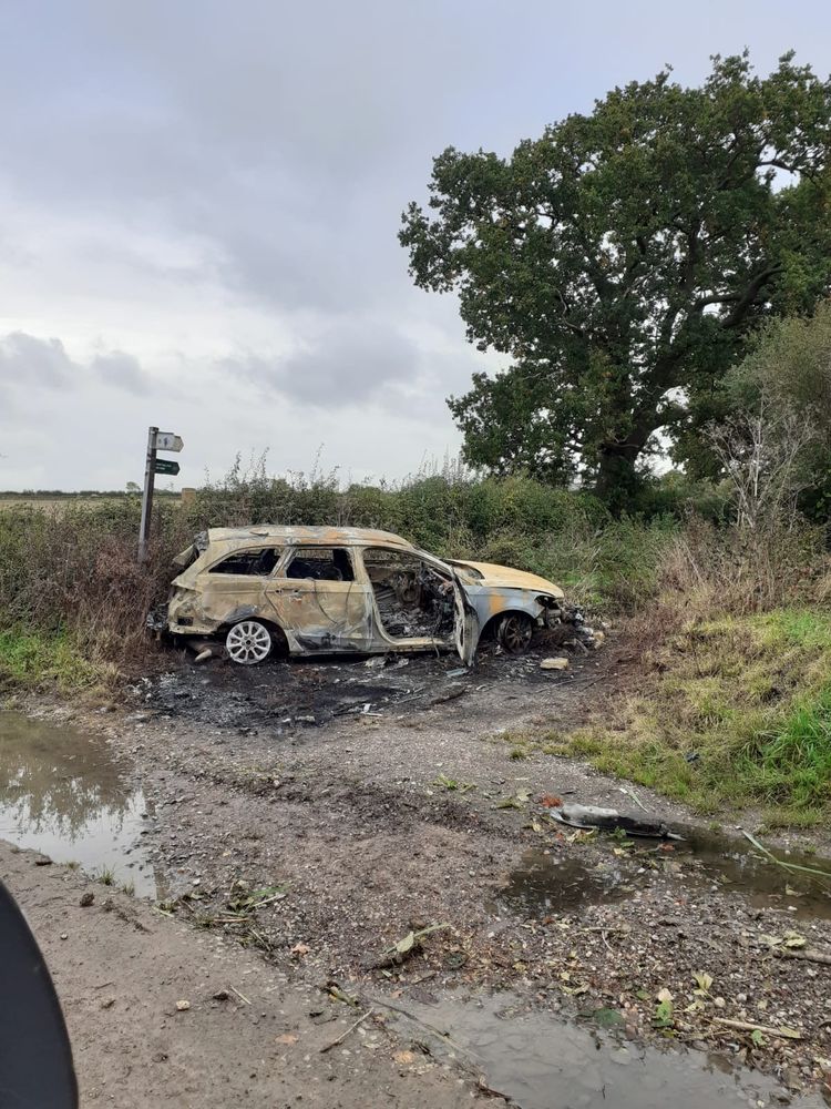The image shows a severely burned-out car, left abandoned on a rural roadside. The vehicle's exterior is charred, and the front end seems particularly damaged. It is positioned near overgrown vegetation and a large tree, with a muddy patch in the foreground. There is also a small signpost in the background, which appears to mark a footpath or country road, though it is partially obscured. The scene conveys a desolate and somewhat neglected environment.


