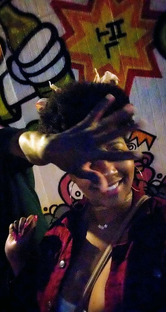 A black woman poses in front of a graffitied wall at night wearing an afro hair style, dark pink denim blazer and crossbody strap across her chest. A dark skinned man's hand blurs as he reaches to cover her face right as she smiles for a selfie so that only part of her face is visible. Her eyes are covered and you can only make out her cheeks rounded in a smile and her nose.