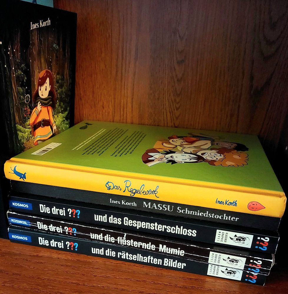 A stack of comic books by Ines Korth: Das Regelwerk, Massu Schmiedstochter, Die drei Fragezeichen und das Gespensterschloss, Die drei Fragezeichen und die flüsternde Mumie, Die drei Fragezeichen und die rätselhaften Bilder 
