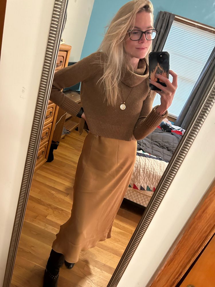 Blonde woman standing in front of a mirror taking a mirror selfie. She’s wearing a gold midi dress, tan cropped cowl neck sweater, black leather boots, and white pendant necklace. 