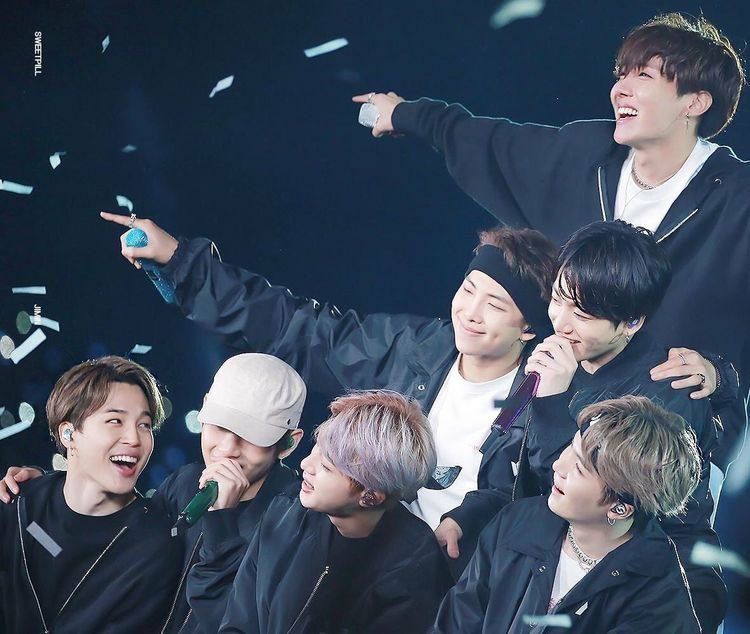 7 members of BTS, all dressed in black hoodies huddled together on stage singing. A photo taken from one of the LY tour stops. There's white confetti falling around them as they look joyous. 