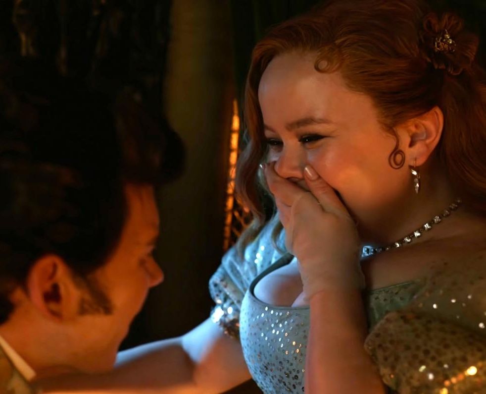 Colin Bridgerton and Penelope Featherington. Penelope is covering her smile with a grin as she stares at Colin who is giggling back with her. 