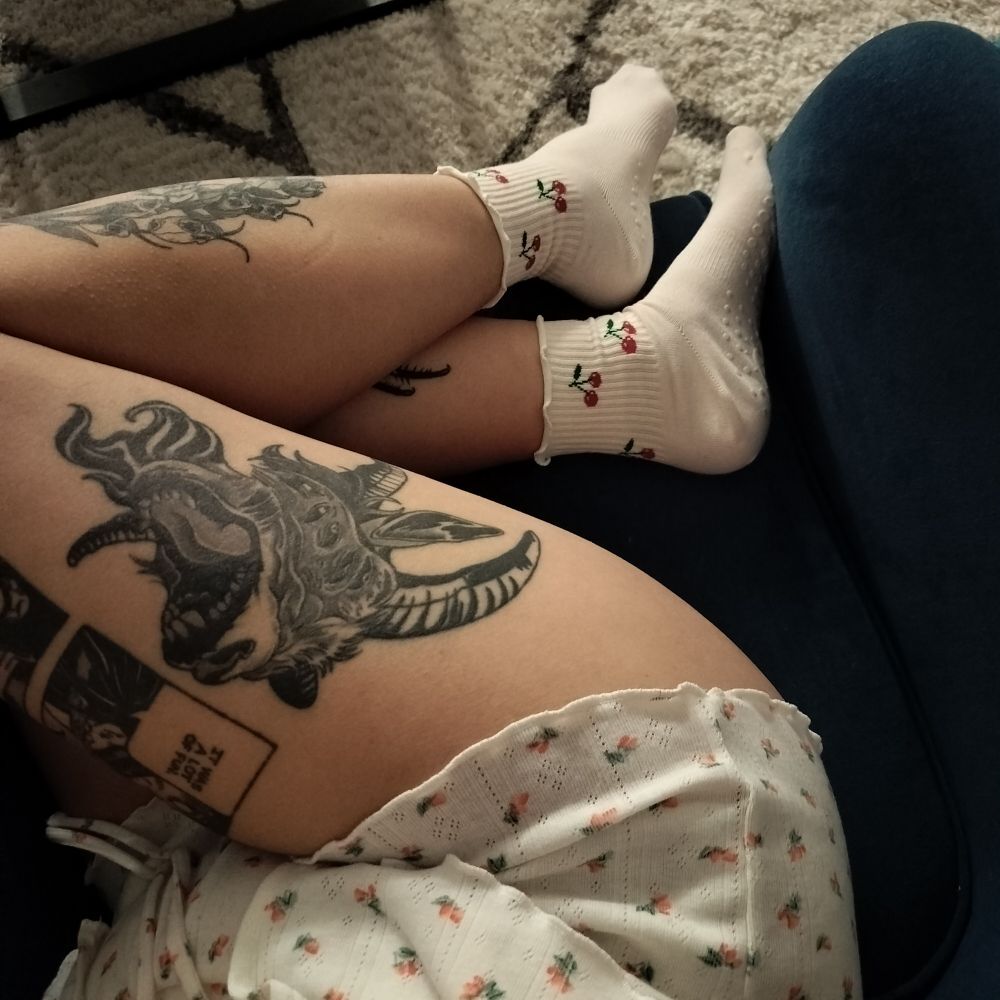 A picture of my legs as I'm sitting on a blue chair. I am wearing white and pink pajamas and white socks with cherries. A goat tattoo on my leg is visible. 