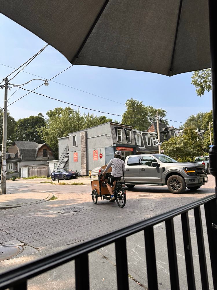 In foreground is a cargo bike - man biking, child in front under an umbrella. In background is an oversized truck. On a residential street. 
