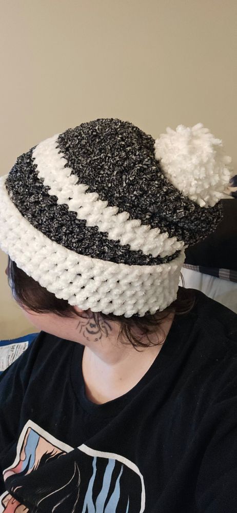 OP shown facing away from camera wearing a hand crocheted beanie in mottled grey and white bold stripe. It has a fluffy white pompom, then a bold grey stripe, a thinner white stripe, then another bold grey stripe, and the cuff is a white stripe. 