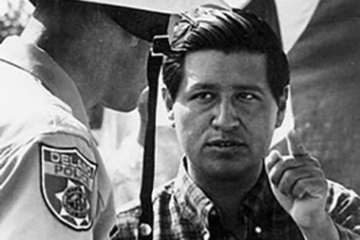 Black and white photo of Cesar Chavez speaking to a Delano police officer whose face is not visible. Chavez's hand is raised and he is pointing for emphasis. Photo from the Cesar Chavez Foundation website.