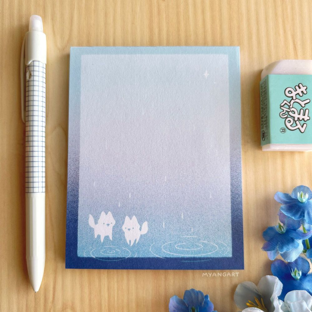 A notepad with an illustration of two white foxes in the bottom left corner, looking at rain puddles drawn with white lines. The background is a gradient from light blue at the top to medium blue at the bottom. It has a dark blue border that also has a gradient going from light to dark from top to bottom. A pen lies to the left of the notepad and an eraser lies to the right. A small bunch of blue flowers borders the lower right hand corner of the picture. Everything lies on top of a light wooden background. 