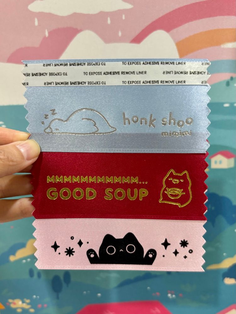 Three badge ribbons. The first is light blue and has the text “honk shoo mimimi,” with an image of a goose sleeping. The second is red with the text “MMMMMMMM… GOOD SOUP” with a drawing of a cat holding a bowl of soup. The third is pink with a drawing of a black cat holding its paws up, with stars on both sides. 