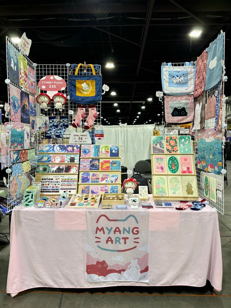 Photo of my artist alley table display, featuring art prints, stickers, tote bags, plushes, charms, and more. There are two grid walls with merchandise, and everything is on top of a pink tablecloth. 