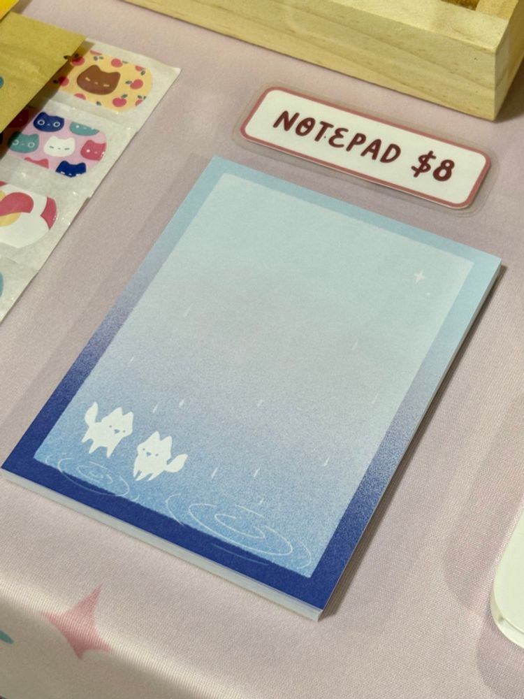Notepad with artwork in the bottom left corner of two white foxes standing in the rain, with puddles at their feet. There is a gradient from medium to light blue going from the bottom to the pad. There is a border, which also has a gradient from dark to light blue. 
