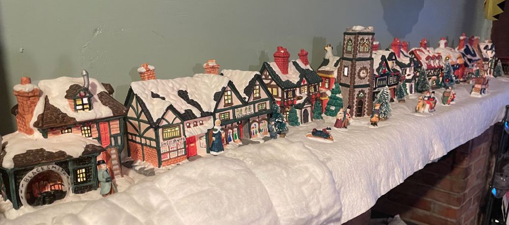 A row of Christmas decorations in the form of ceramic buildings, sitting on pretend snow. Nearest to us is the Blacksmiths, with its open fire, then a row of Shoppes, then it gets harder to tell what the buildings are, except for the grey church with its tall Norman tower. There are also trees and figures.