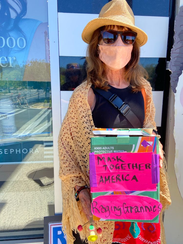 Granny Linda, wearing light orange KF94 mask, held a box of KN95 masks that was covered with a sign that says Mask Together America and Raging Grannies