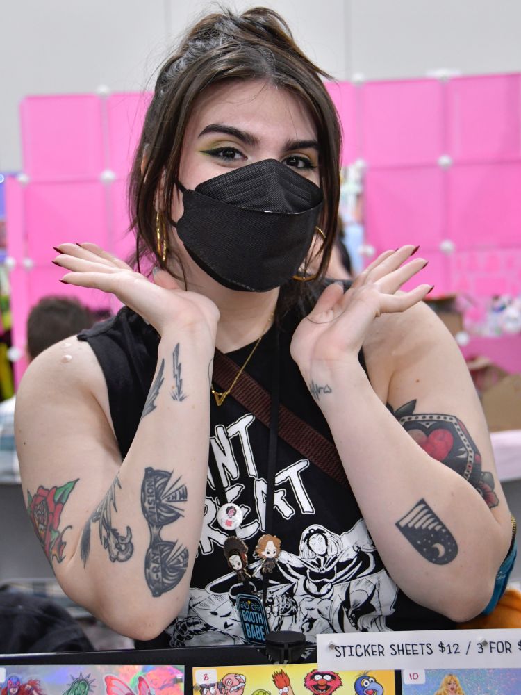 Alex Lajas, wearing a black KN95 respirator, black T-shirt with graphic and text that says, “Mutant Threat,” tabling for Lauren Moran at New York Comic Con, posed for the pandemic photographic documentary project Julie Lam created for MaskTogetherAmerica. 