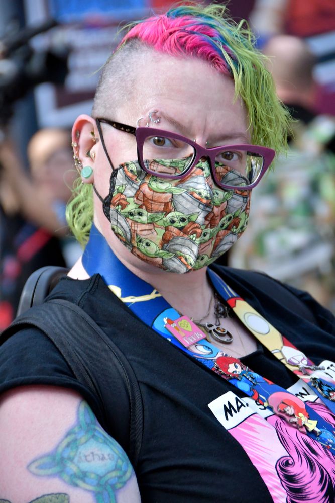 Karen Walsh, double-masking with a black KN95 under a cloth mask made with pattern of Grogu (Baby Yoda) from the Mandalorian. Karen was wearing a pair of purple framed glasses, a black top with graphics on it. She also has multiple piercing on her eyebrows, nose and ears, multi-color mohawk hairstyle, and tattoos on her arm.
