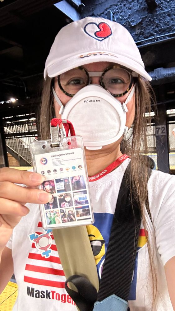 Julie, wearing a flo mask, a hat with IgAN Foundation’s logo, a team T-shirt of MaskTogetherAmerica with a sticker to identify her support for the Transgender community, carrying heavy equipment in a camera bag, on her way to the Pride Health Fair in 2024.