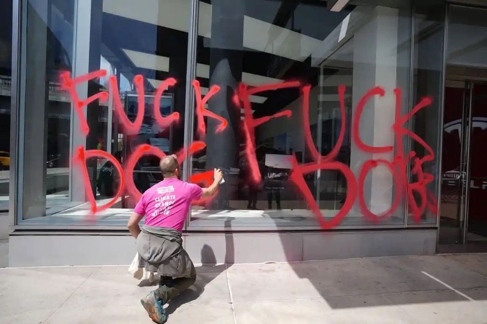 Tesla dealership with XR NYC spraying on the words 'Fuck Doge' with spray chalk.