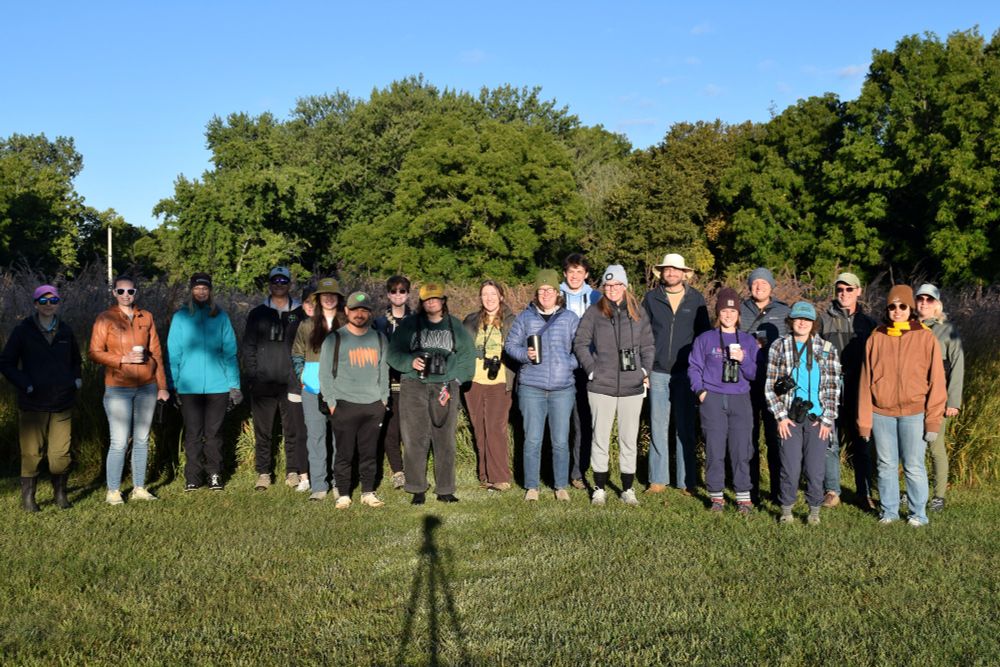 A group of people smiling and holding binoculars getting ready to go birding. The park has a lawn and trees in the background.