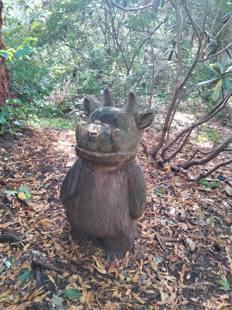 Wooden carving of a creature, I think it's a baby gruffallo in a woodland setting. Leaves on the ground and dappled autumn sunlight 