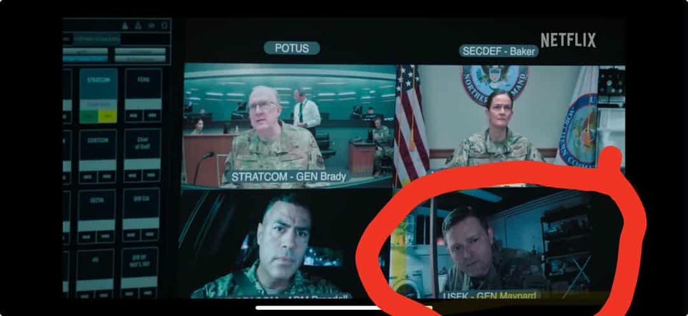 Screenshot depicting a video conference call with several military officers participating from watch floors and conference rooms. One officer is crouched in a dim basement. In the background is what looks like an LG or Samsung front-loading washing machine, utility shelves, and a pole. 