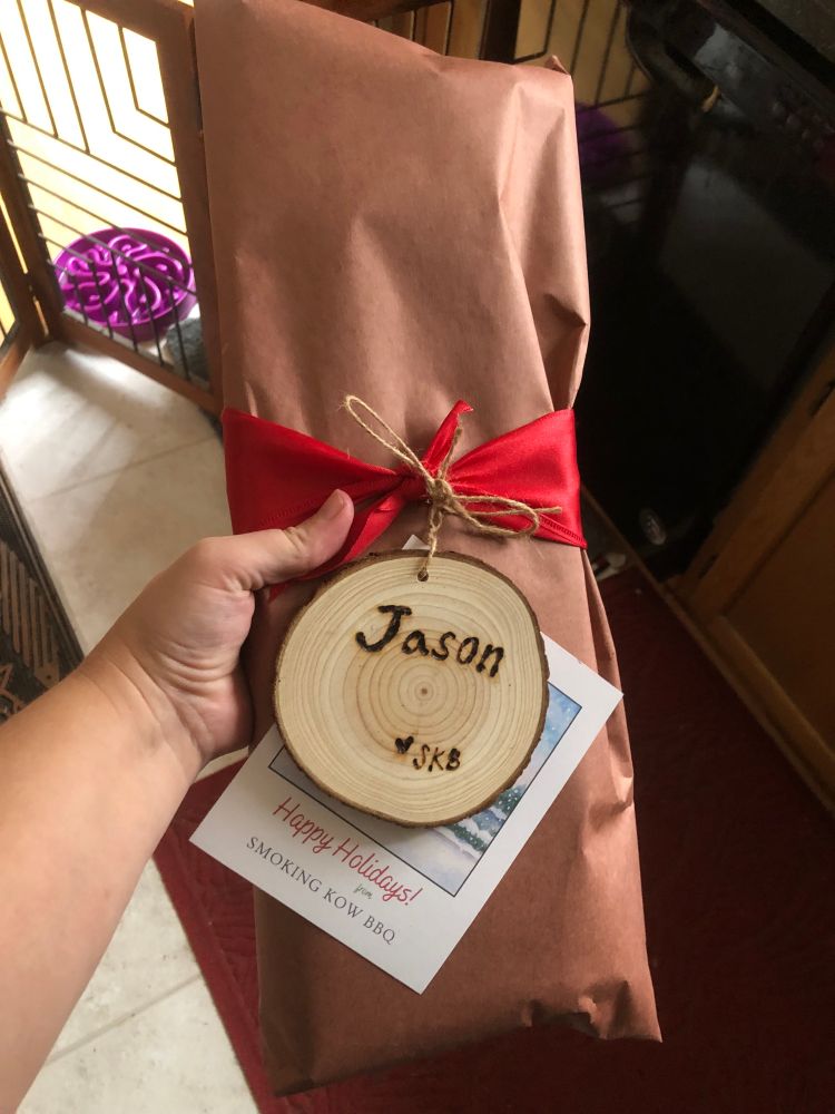 A wrapped parcel of brown butcher paper tied with a red ribbon and a gift tag made out of a round piece of wood with the name "Jason" carved (or burnt?) into it, being held up in front of the camera. A card underneath the tag reads "Happy Holidays from Smoking Kow BBQ."