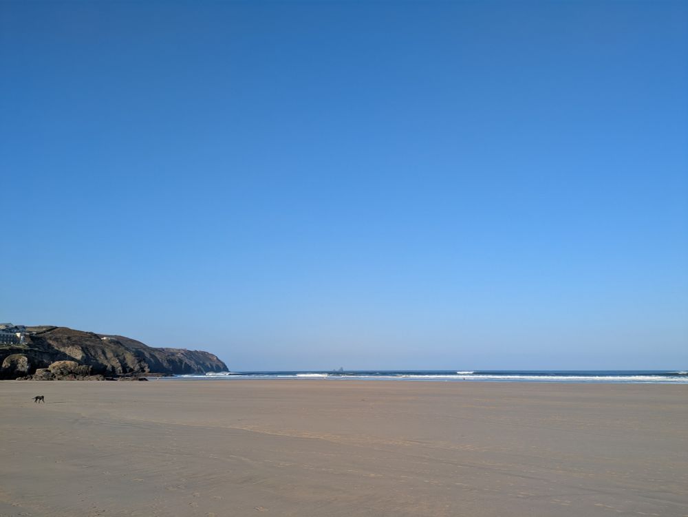 Perranporth beach. Cliffs on the left, an expanse of empty yellow sand and cloudless blue sky above. Hint of crashing waves separates the horizon.