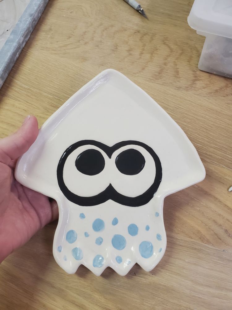 An approx 10 inch white stoneware plate in the shape of the squid icon from splatoon with blue spots at the bottom