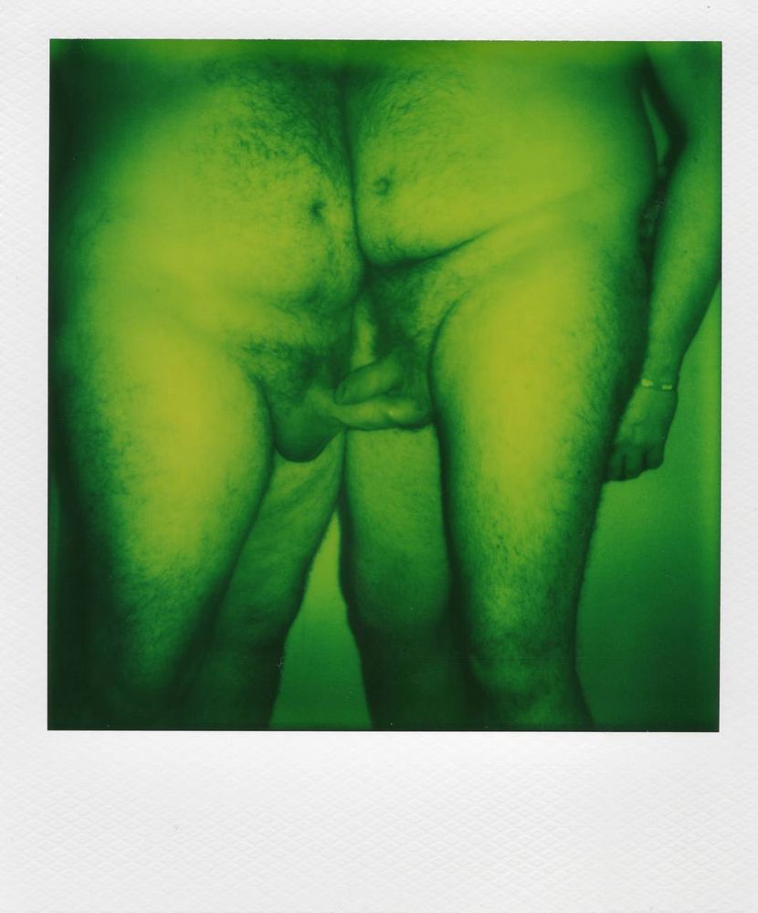 A scan of a Polaroid photograph showing two people nude stood facing each other, the camera level with their crotch. The figure on the left is erect, and the figure on the right’s flaccid cock is resting on it. The film is monochrome, but with a scale of green to black 
