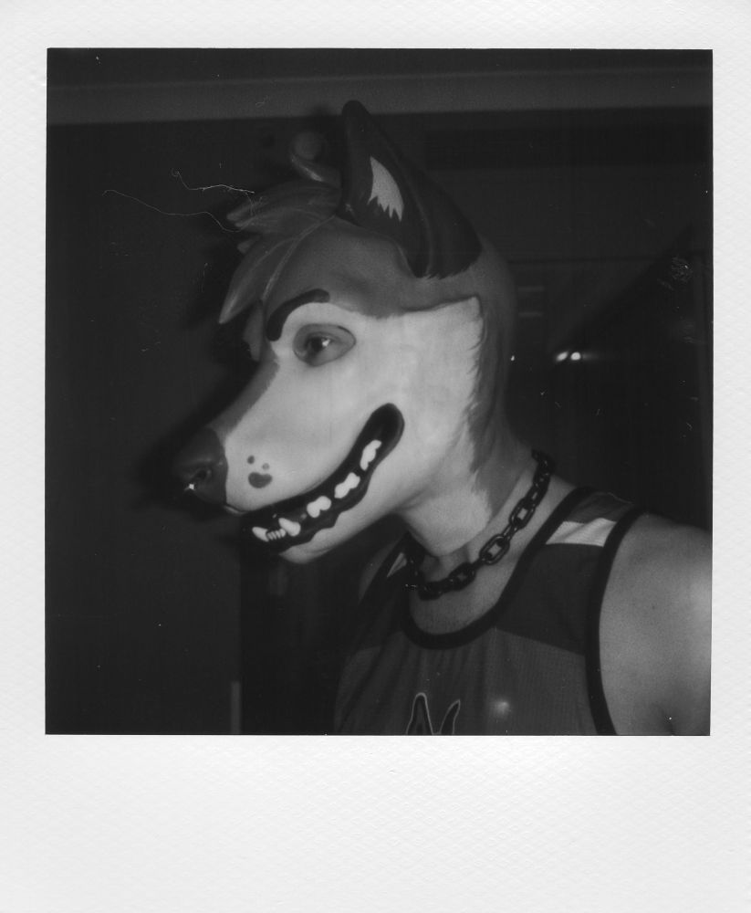 A black and white Polaroid photograph of a person in a tank top and a rubber dog hood of their fursona. They look cute, tell them they’re good 
