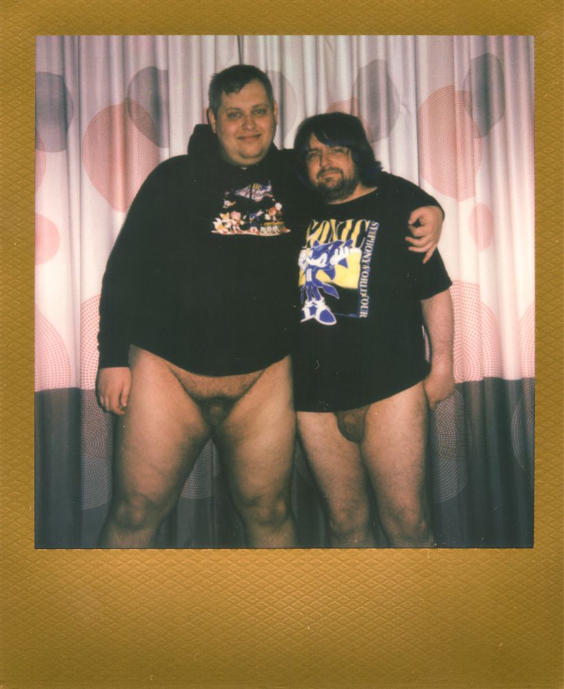 A Polaroid photograph with a gold frame. The photo shows my friend in the left and me on the right, his arm is on my shoulder. We’re both wearing band shirts for the show we’re going to. Also, we’re naked from the waist down and you can see our flaccid uncut cocks