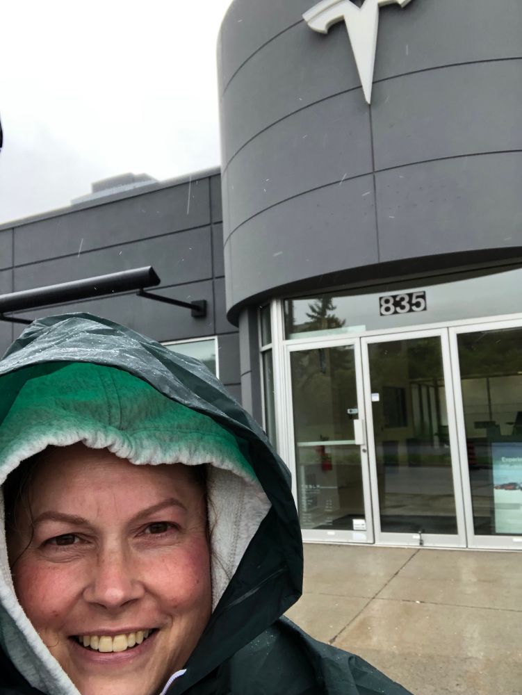 Smiling woman in a grey hoodie and dark green poncho standing in front of a Tesla dealership in the rain. 