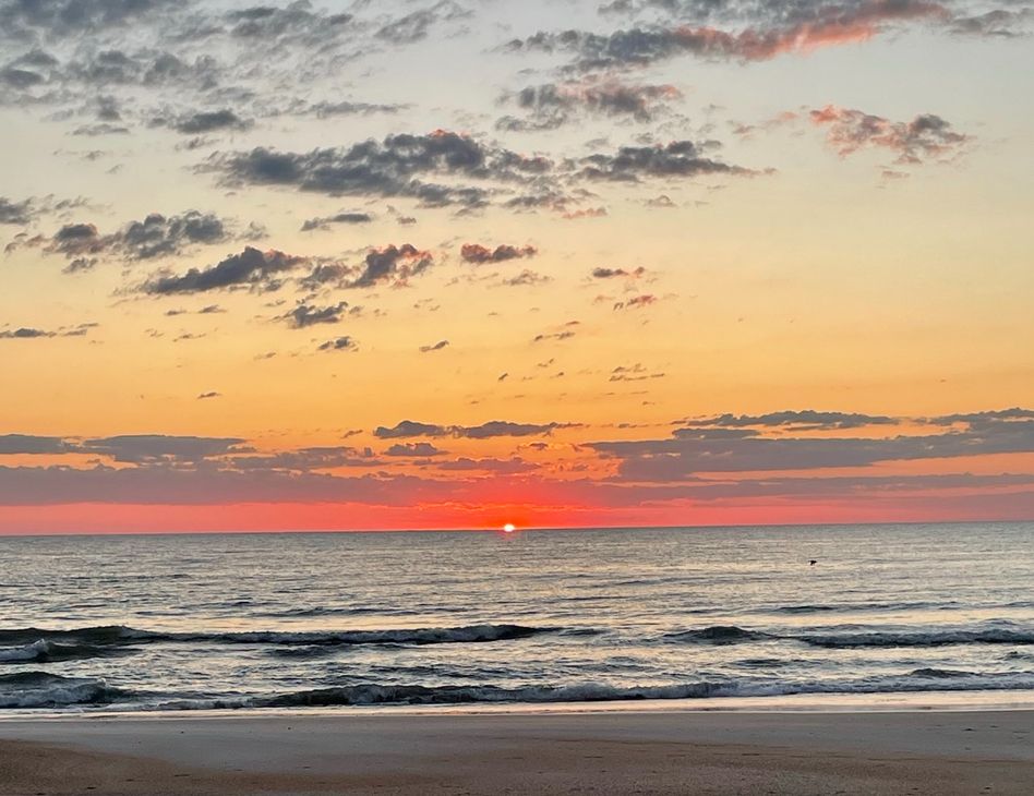 The sun has just crested over the ocean reflecting orange in the sky with slight cloud coverage above. The ocean with very small waves hitting the beach is in the foreground.