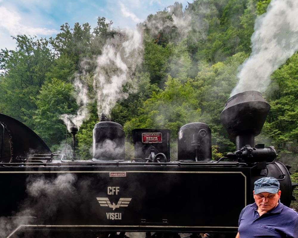 A close-up, low-angle shot of the black steam locomotive of the Mocănița narrow-gauge railway in Viseu de Sus, Romania. Plumes of thick, white steam billow upwards from the chimneys and around the body of the train, obscuring the lush green forest hillside in the background. The text 'CFF VISEU' and the stylized wing logo are visible on the side of the engine. A man in a blue shirt and cap is partially visible in the bottom right corner.