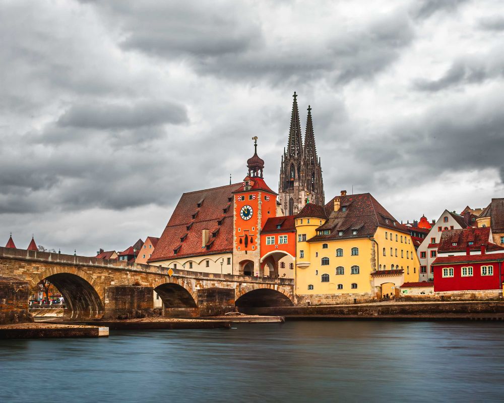A vibrant, long-exposure photograph of Regensburg, Germany, focusing on the Stone Bridge crossing the Danube River. In the center, the bright orange-red roof and clock tower of the Old Town Hall stand out against a dramatic, heavily clouded sky. Behind it, the tall, twin spires of the Regensburg Cathedral are visible. The river is a smooth, deep blue due to the long exposure, and colorful historic buildings (yellow, red, orange) line the right bank