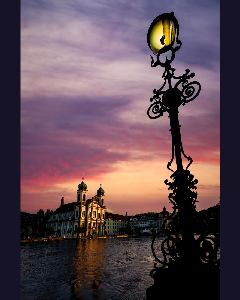 A dramatic vertical image of the Jesuit Church in Lucerne, Switzerland, illuminated by lights at the water's edge. The church is framed on the right by a large, ornate, black iron lamp post in silhouette. The sky above is a vibrant blend of deep pink, purple, and orange from the sunset, reflecting faintly on the flowing water of the Reuss River.
