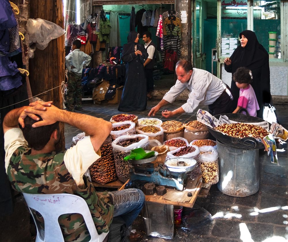 A street-level, medium shot taken inside a bustling covered market in Damascus. In the foreground on the left, a man wearing a camouflage vest and jeans sits in a white plastic chair with his back to the camera, resting his hands behind his head. To his right is a dense display of burlap sacks and metal bowls filled with various roasted nuts and seeds, sitting next to an old-fashioned blue mechanical scale.
In the mid-ground, a customer with a receding hairline and a white shirt leans over the display, reaching toward the goods. To the right, a woman wearing a full-length black abaya and head covering stands near the stall with a small child in a pink shirt. In the background, hanging clothes line the stone-paved alleyway, and other pedestrians can be seen walking through the market in the distance.
