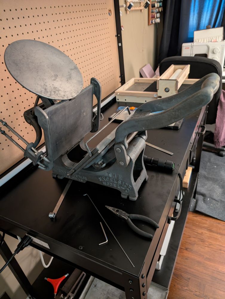 A 5x8" Kelsey press, a Provisional Press, and a very shitty paper cutter on a black workbench with a pegboard. Sewing machine visible in the back, quoin key, composing sticks, and galleys visible on the bottom shelf of the workbench.