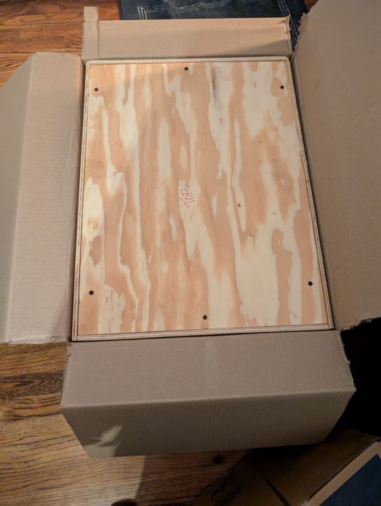 Top of a plywood crate inside a cardboard box. Top written in red marker.
